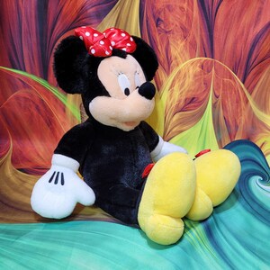 K&ouml;nnte beinhalten: Eine Pl&uuml;schpuppe von Minnie Mouse, mit einem schwarzen K&ouml;rper, wei&szlig;en Handschuhen und gro&szlig;en gelben F&uuml;&szlig;en. Sie tr&auml;gt eine rote Schleife mit wei&szlig;en Punkten. Die Puppe sitzt vor einem bunten, abstrakten Hintergrund.