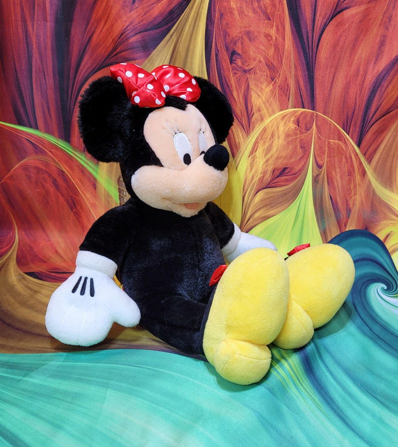 K&ouml;nnte beinhalten: Eine Pl&uuml;schpuppe von Minnie Mouse, mit einem schwarzen K&ouml;rper, wei&szlig;en Handschuhen und gro&szlig;en gelben F&uuml;&szlig;en. Sie tr&auml;gt eine rote Schleife mit wei&szlig;en Punkten. Die Puppe sitzt vor einem bunten, abstrakten Hintergrund.