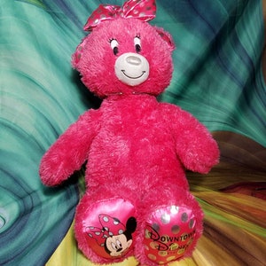 May include: A bright pink plush teddy bear with a matching bow featuring silver polka dots. The bear has a smiling face and the Downtown Disney District logo on its foot. Minnie Mouse is also featured on the foot.