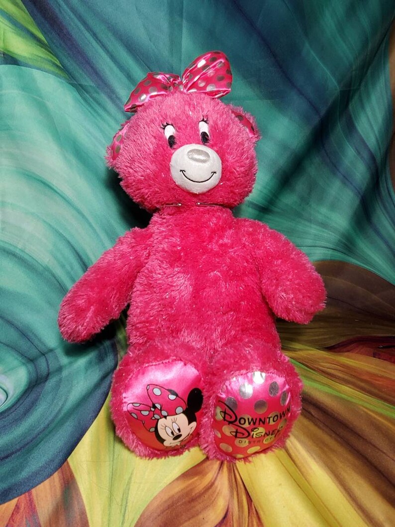 May include: A bright pink plush teddy bear with a matching bow featuring silver polka dots. The bear has a smiling face and the Downtown Disney District logo on its foot. Minnie Mouse is also featured on the foot.