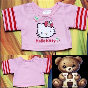 Może przedstawiać: R&oacute;żowa koszulka z czerwono-białymi paskami na rękawach. Z przodu grafika Hello Kitty, z jej twarzą, czerwoną kokardą i napisem "Hello Kitty". Wiśnie dopełniają projekt.