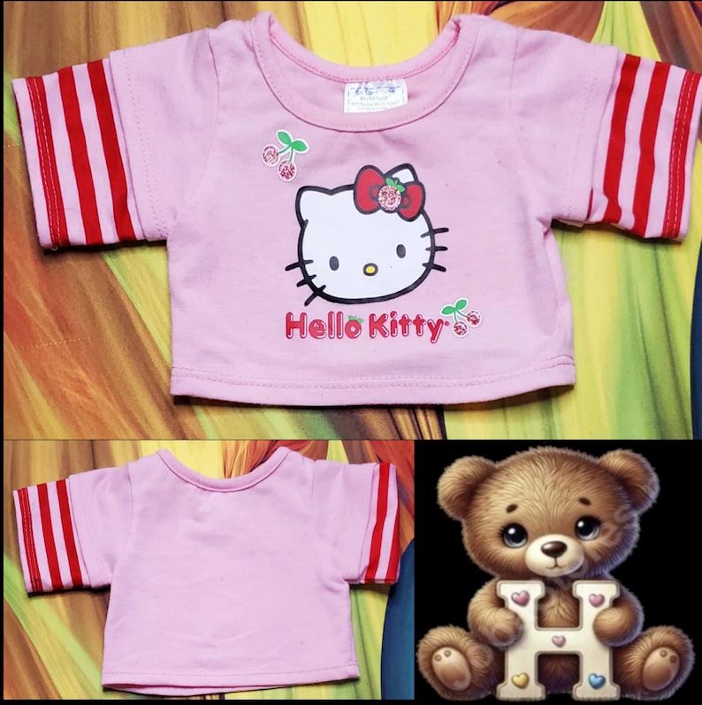 Może przedstawiać: R&oacute;żowa koszulka z czerwono-białymi paskami na rękawach. Z przodu grafika Hello Kitty, z jej twarzą, czerwoną kokardą i napisem "Hello Kitty". Wiśnie dopełniają projekt.