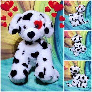 May include: A white and black dalmatian plush dog with a red heart on one eye. The dog is sitting with its legs crossed and has a playful expression.