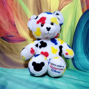 Pu&ograve; includere: Un orsacchiotto di peluche bianco con macchie colorate rosse, gialle, blu e nere. L'orso ha un naso nero e occhi ricamati. Il piede destro ha il testo "Custom made at: Downtown Disney". Il piede sinistro ha una testa di Mickey Mouse nera.