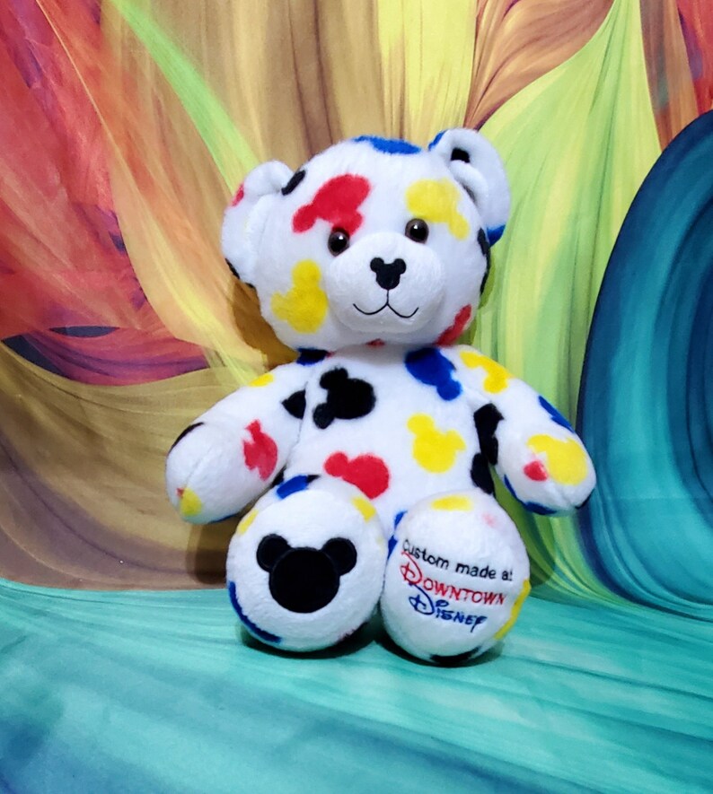 Pu&ograve; includere: Un orsacchiotto di peluche bianco con macchie colorate rosse, gialle, blu e nere. L'orso ha un naso nero e occhi ricamati. Il piede destro ha il testo "Custom made at: Downtown Disney". Il piede sinistro ha una testa di Mickey Mouse nera.