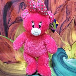 Puede incluir: Un oso de peluche rosa brillante con un lazo a juego con lunares blancos y verde claro. El oso tiene un hocico blanco, pesta&ntilde;as negras y una etiqueta. El juguete est&aacute; frente a un fondo colorido y abstracto.
