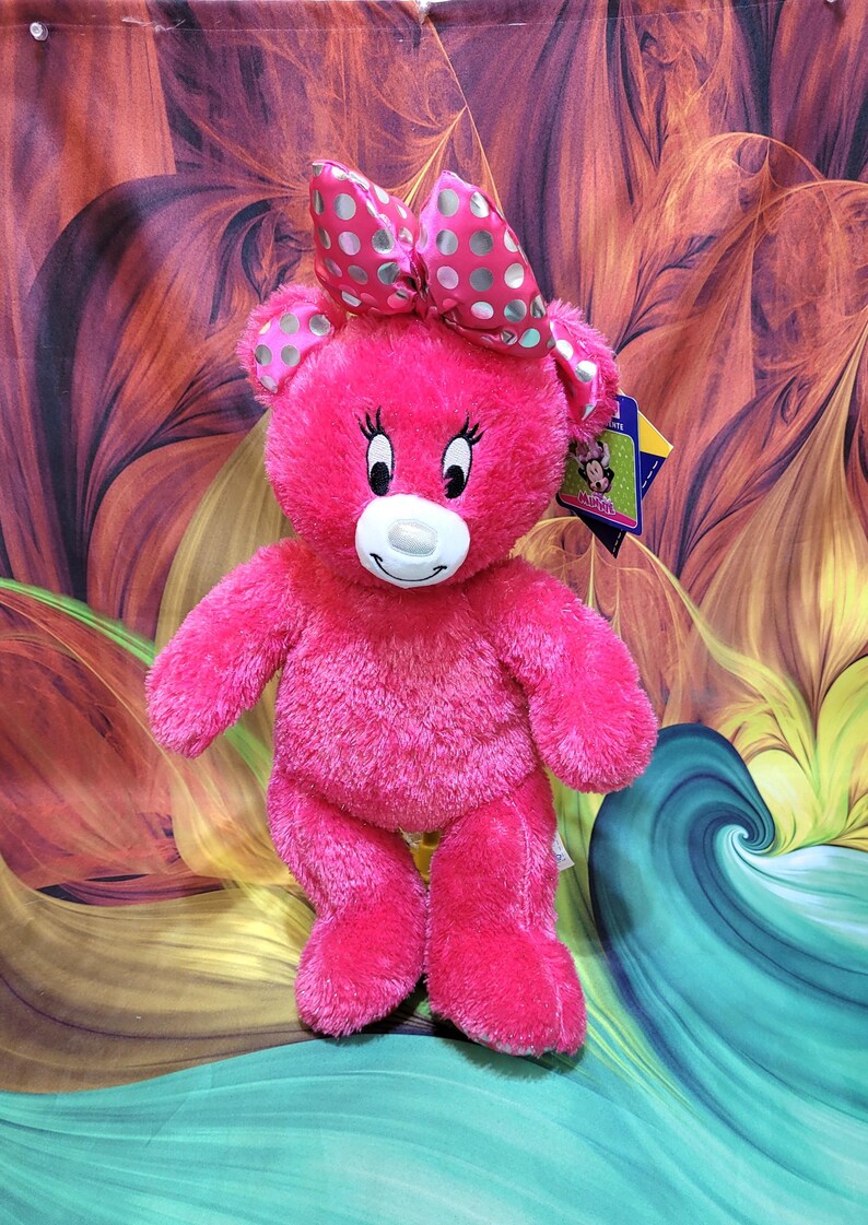 Puede incluir: Un oso de peluche rosa brillante con un lazo a juego con lunares blancos y verde claro. El oso tiene un hocico blanco, pesta&ntilde;as negras y una etiqueta. El juguete est&aacute; frente a un fondo colorido y abstracto.