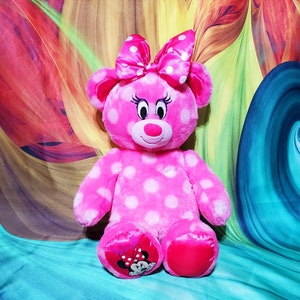 Op de afbeelding: Een roze pluche beer met witte stippen, een bijpassende strik en Minnie Mouse-afbeeldingen op de voeten. De beer heeft een lachend gezicht met wimpers en een rode neus. De achtergrond heeft een kleurrijk, abstract ontwerp.