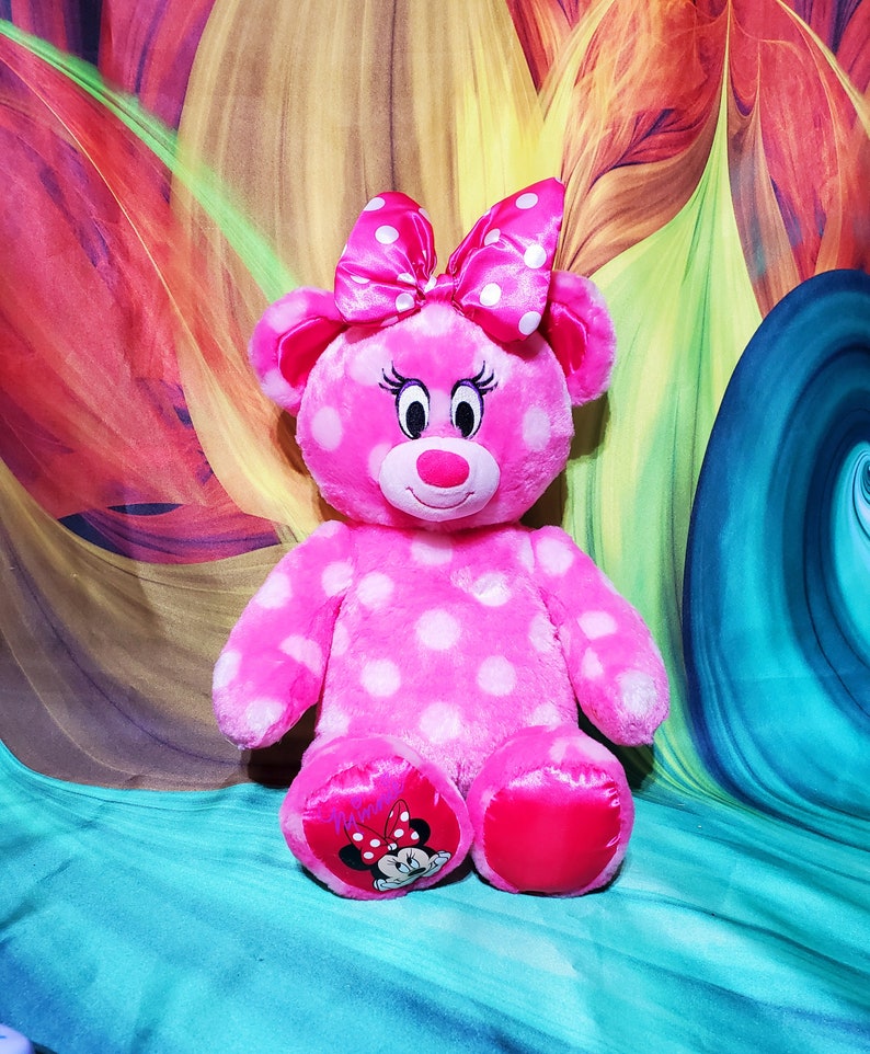 Op de afbeelding: Een roze pluche beer met witte stippen, een bijpassende strik en Minnie Mouse-afbeeldingen op de voeten. De beer heeft een lachend gezicht met wimpers en een rode neus. De achtergrond heeft een kleurrijk, abstract ontwerp.