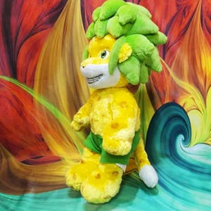 May include: A plush toy lion with a yellow body and green mane and shorts. The lion has a smiling face with white teeth and paws. The background is a colorful abstract design with swirls of red, orange, and blue.