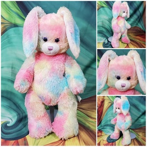 May include: A plush bunny rabbit with long ears and a tie-dye pattern in shades of pink, blue, and yellow. The stuffed animal has a pink nose, dark eyes, and a soft, fluffy texture. The bunny is standing upright.