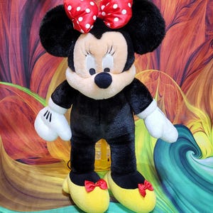 K&ouml;nnte beinhalten: Eine Pl&uuml;schpuppe von Minnie Mouse, mit einem schwarzen K&ouml;rper, wei&szlig;en Handschuhen und gelben Schuhen mit roten Schleifen. Die Puppe hat ein cremefarbenes Gesicht, schwarze Ohren und eine rote Schleife mit Punkten. Das Spielzeug ist dem ikonischen Disney-Charakter nachempfunden.
