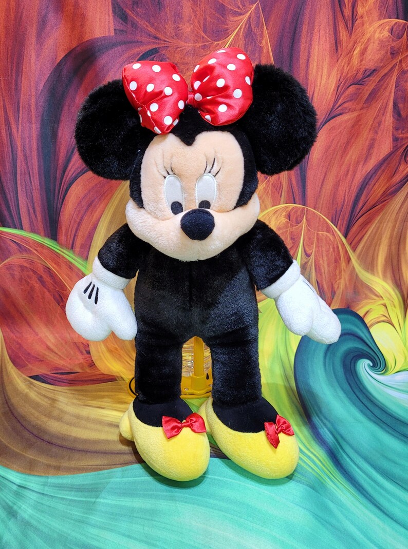K&ouml;nnte beinhalten: Eine Pl&uuml;schpuppe von Minnie Mouse, mit einem schwarzen K&ouml;rper, wei&szlig;en Handschuhen und gelben Schuhen mit roten Schleifen. Die Puppe hat ein cremefarbenes Gesicht, schwarze Ohren und eine rote Schleife mit Punkten. Das Spielzeug ist dem ikonischen Disney-Charakter nachempfunden.
