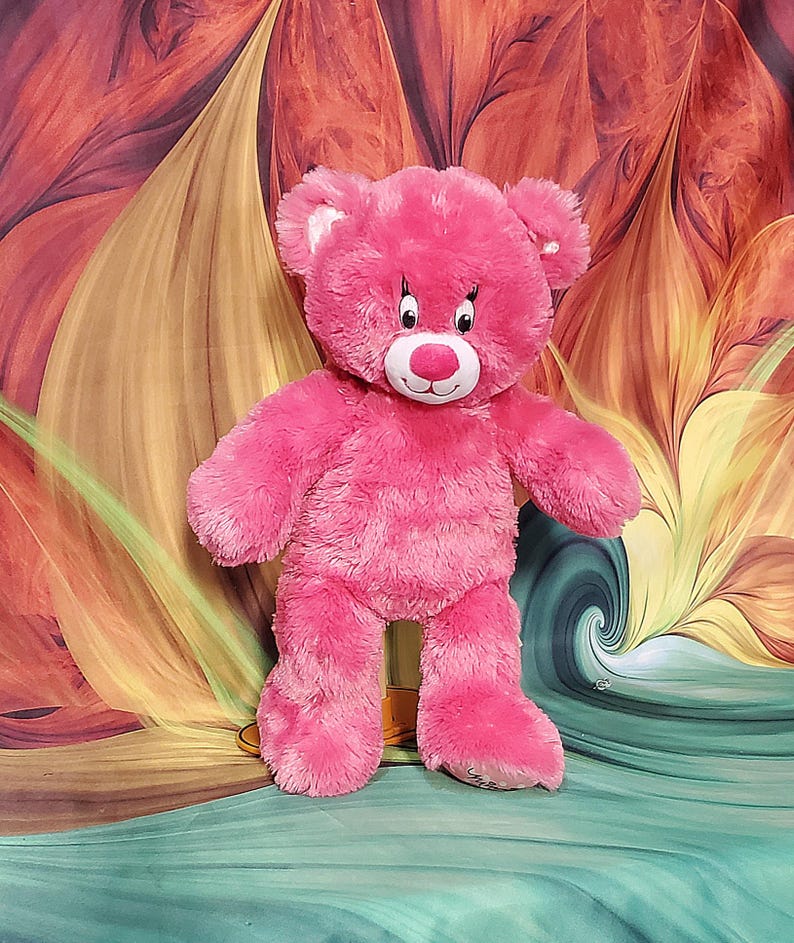 K&ouml;nnte beinhalten: Ein rosa Teddyb&auml;r mit wei&szlig;er Schnauze und Ohren, schwarzen Augen und einer roten Nase. Der B&auml;r steht aufrecht mit ausgestreckten Armen und Beinen. Der Hintergrund zeigt ein farbenfrohes Blumendesign mit Rot-, Orange- und Gelbt&ouml;nen.