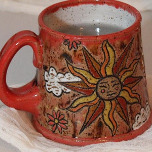 May include: A red ceramic mug with a hand-painted design of a sun with a face and clouds. The mug is sitting on a white cloth next to a silver teapot.