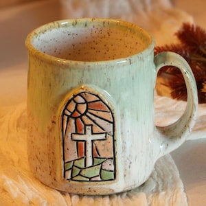 May include: Stack of three green ceramic mugs with a stained glass design featuring a cross. The mugs are stacked on a light brown surface. A single mug with the same design is on a white fabric surface.