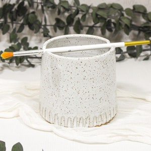 May include: White ceramic brush holder with a speckled finish and a white paintbrush inside. The holder has a decorative scalloped edge.