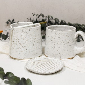 May include: Two white ceramic mugs with a speckled glaze and a white ceramic dish with a textured design. The mugs have a rounded shape and a handle. The dish is round and has a slightly raised edge.