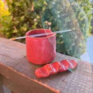 May include: A red ceramic paint brush holder with a brush resting on the rim. A matching red ceramic paint palette knife is next to the holder.