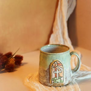 May include: A ceramic mug with a speckled light green and tan glaze, featuring a raised, arched emblem on the front. The emblem depicts a cross set against a sunburst pattern in shades of orange, yellow, and green, resembling a stained glass window.