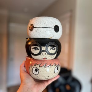 May include: A stack of three ceramic bowls, each hand-painted with a cartoon character design. The top bowl is white with black accents, the middle is black with glasses, and the bottom is peach with red hair.