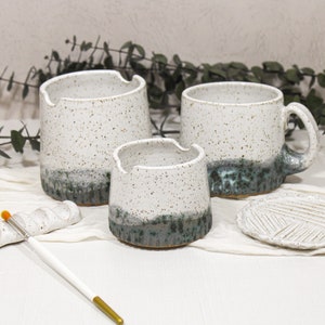 Puede incluir: Tres tazas de cerámica blanca con un esmalte moteado y una banda verde azulada alrededor de la base. Las tazas están dispuestas sobre una superficie blanca con un portacepillos de cerámica blanca y un pincel.