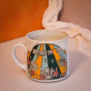 May include: A white ceramic mug with a speckled glaze. The mug features a colourful design of a tent with a blue, yellow and green striped awning. The tent is surrounded by white flowers with pink centres.