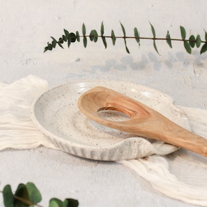 May include: White ceramic spoon rest with a speckled glaze and a wooden spoon resting in the holder.