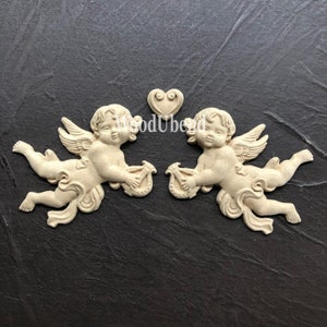 May include: Two wooden cherub figurines holding musical instruments, with a heart-shaped design above them. The cherubs are painted in a light beige color and have delicate wings.