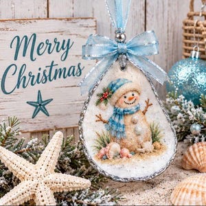 Può includere: Un ornamento a forma di goccia ricavato da una conchiglia di ostrica con un pupazzo di neve dipinto che indossa sciarpa e cappello blu, sormontato da un fiocco di nastro azzurro. La conchiglia è decorata con sabbia e piccole conchiglie.