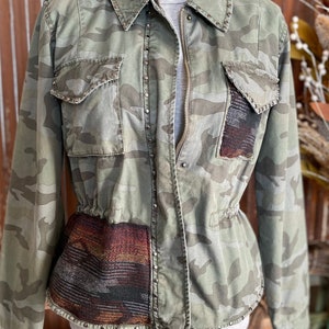 May include: A green camouflage jacket with a zipper closure and a brown and black patterned fabric panel on the bottom. The jacket has silver studs around the edges and pockets.