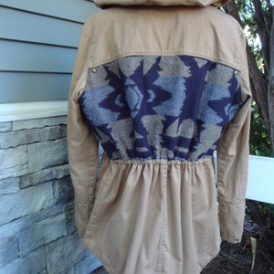 May include: A tan hooded jacket with a blue and brown geometric pattern on the back. The jacket has a drawstring waist and a long, flowing style.