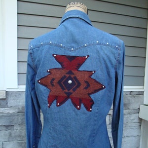 May include: A blue denim shirt with a brown and red geometric pattern on the back. The pattern is made of fabric and has a dark blue center.