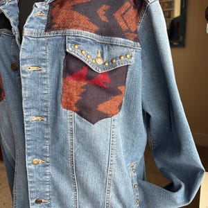 May include: A blue denim jacket with a southwestern patterned fabric design on the shoulders and pockets. The fabric is a mix of red, orange, and black.