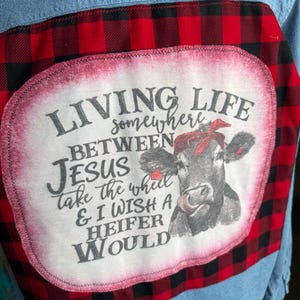 May include: Light blue denim shirt with a red and black plaid back panel. The panel features a white oval patch with the text "LIVING LIFE somewhere BETWEEN JESUS take the wheel & I WISH A HEIFER WOULD" and a cow graphic.