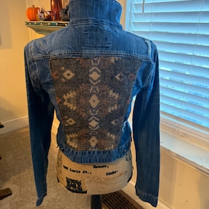May include: A blue denim jacket with a brown and beige patterned fabric patch on the back. The jacket has a distressed look with frayed edges.