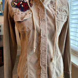 May include: A beige corduroy shirt with a hood and a distressed look. The shirt has a button-up front and a patch on the left chest featuring a vintage-style illustration of three people.