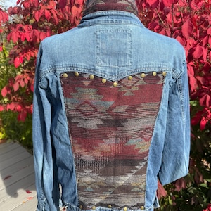 May include: Light blue denim jacket with a patterned fabric panel on the back and collar. The panel features a geometric design in shades of red, brown, and green, accented with brass-colored studs.