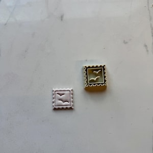 May include: Two white plastic cookie cutters shaped like postage stamps with a design of two birds flying inside. The cookie cutters are on a white marble surface.