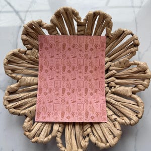 May include: A pink patterned paper with a repeating design of cacti, stars, and other southwestern motifs. The paper is sitting on a woven straw flower-shaped tray.