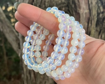 May include: Several white and iridescent beaded bracelets are held in a hand. The bracelets are made of small, round beads. The background is a blurred view of a tree trunk.