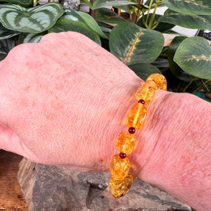 May include: A bracelet featuring amber-colored beads of varying shapes and sizes, interspersed with small, round, dark red beads. The bracelet is displayed on a person's wrist, showcasing its warm, golden hues.