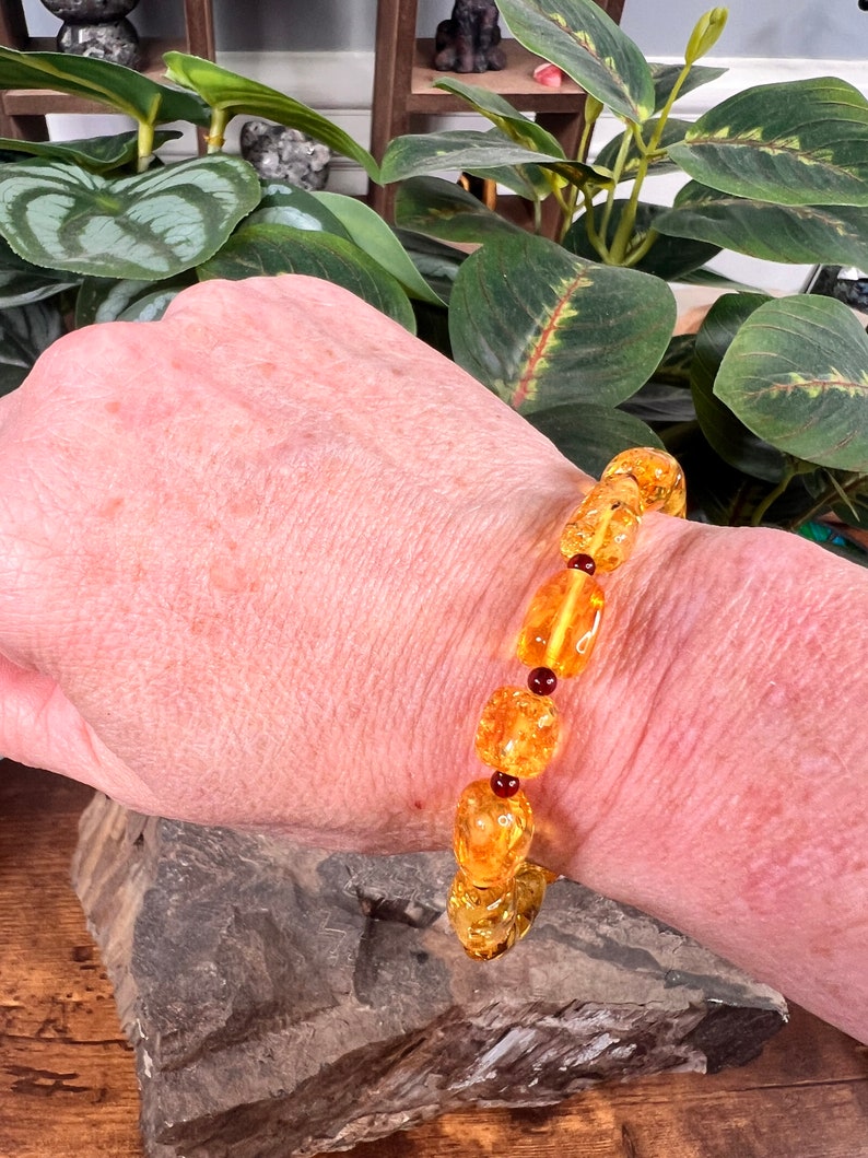May include: A bracelet featuring amber-colored beads of varying shapes and sizes, interspersed with small, round, dark red beads. The bracelet is displayed on a person's wrist, showcasing its warm, golden hues.