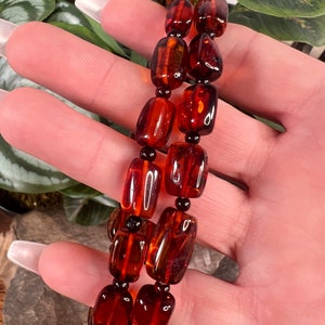 May include: Two strands of amber-colored beads, each with a series of rectangular and round beads. The beads have a glossy finish and are strung together with black beads. The jewelry is displayed against a natural background.