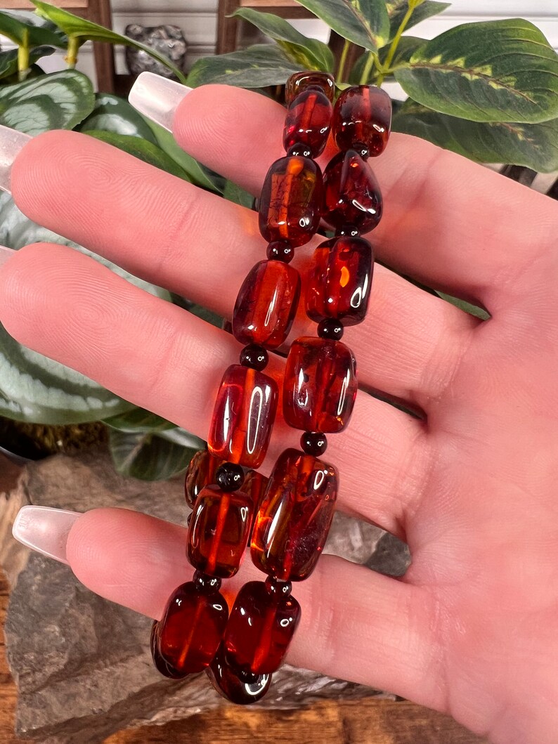 May include: Two strands of amber-colored beads, each with a series of rectangular and round beads. The beads have a glossy finish and are strung together with black beads. The jewelry is displayed against a natural background.