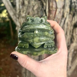 May include: A carved green jade frog figurine. The frog is detailed with textured skin and sits on a leaf-shaped base. The jade has varying shades of green, and the frog is held in a hand.