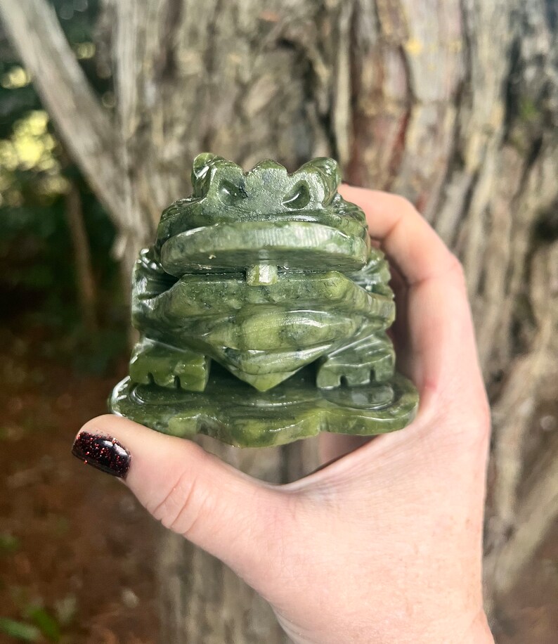 May include: A carved green jade frog figurine. The frog is detailed with textured skin and sits on a leaf-shaped base. The jade has varying shades of green, and the frog is held in a hand.