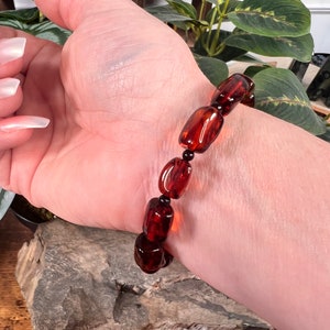 May include: A dark amber bracelet with rectangular beads and small black beads. The bracelet is worn on a wrist, showcasing the rich, translucent color of the amber. The beads are strung together, creating a striking accessory.