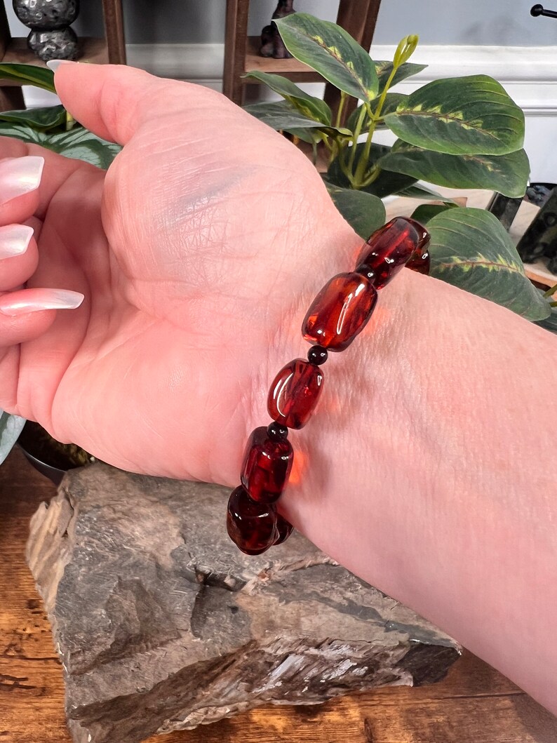 May include: A dark amber bracelet with rectangular beads and small black beads. The bracelet is worn on a wrist, showcasing the rich, translucent color of the amber. The beads are strung together, creating a striking accessory.