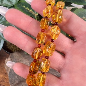 May include: A pair of amber bracelets with a warm, golden hue. The bracelets are made of irregularly shaped amber beads, interspersed with small, dark red beads. The bracelets are displayed against a backdrop of green foliage.